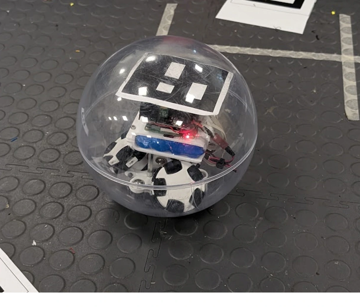 Enclosed sphere robot on arena floor with ArUco marker visible through the acrylic shell